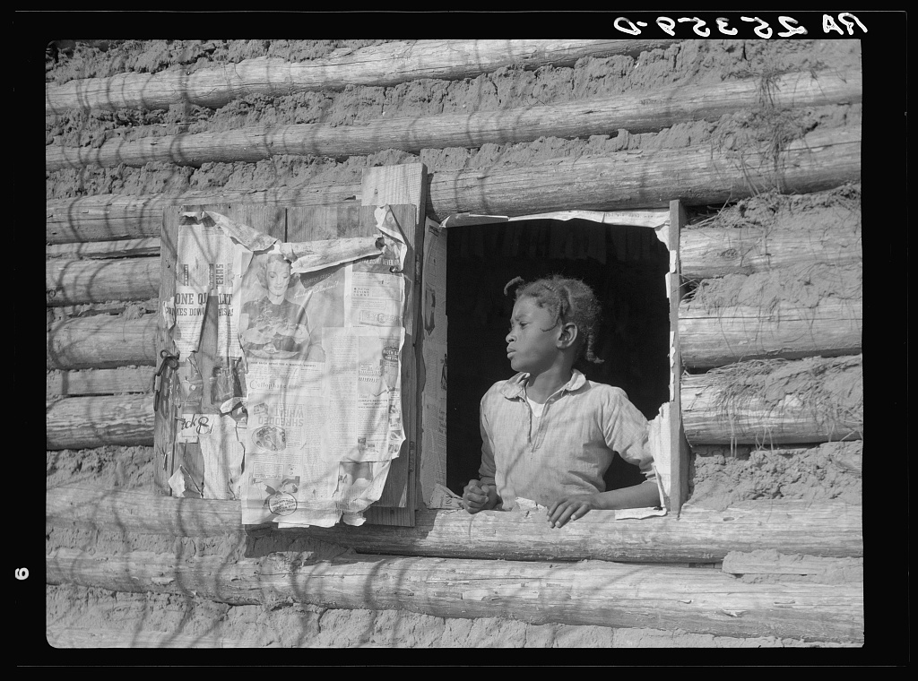 Hair in cornrows, face in gentle repose, a young girl stands at the glassless window of her cabin, a faraway look on her face