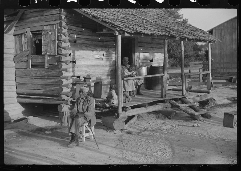 A man sits in a chair on the ground, his back to the porch of an old wooden cabin